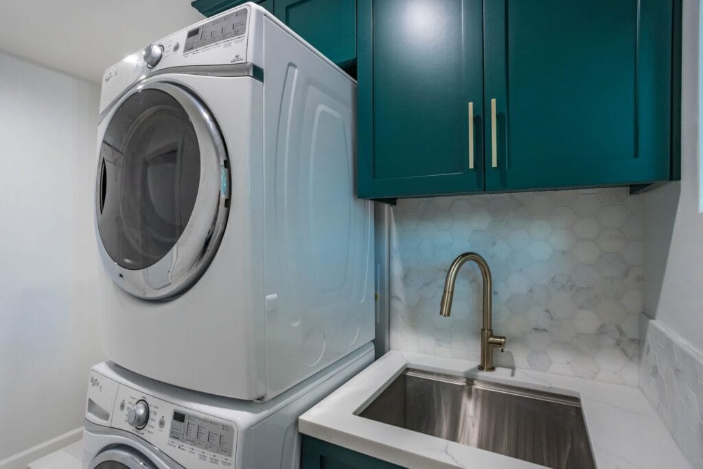 Utility room with a stacked washer and dryer beside a stainless sink set into teal cabinetry under a hexagonal mosaic splashback