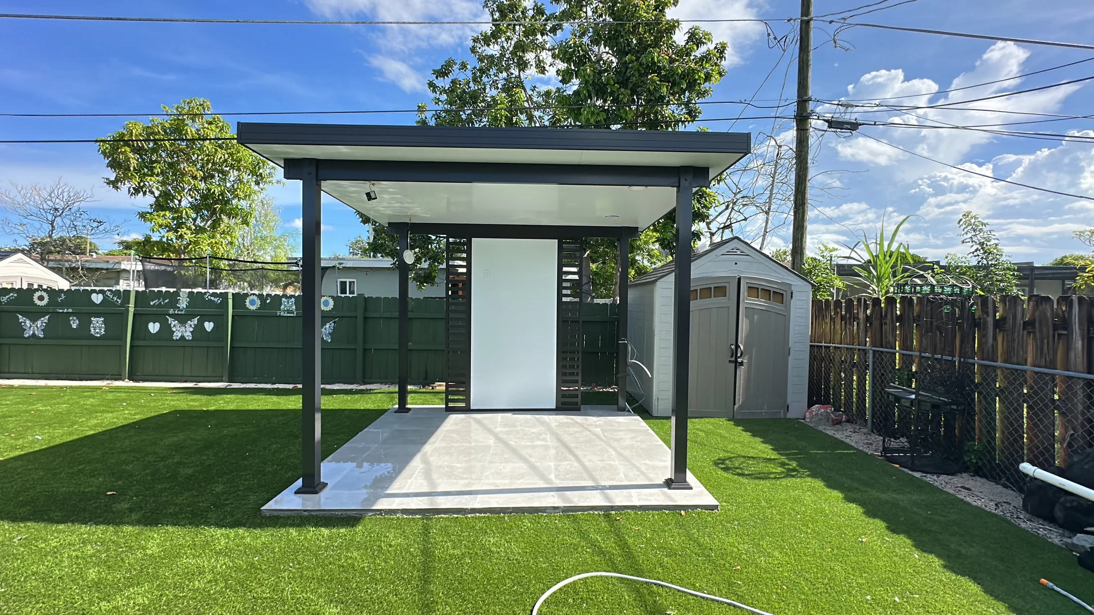 Freestanding pergola with white posts set on a paver pad in a fenced lawn
