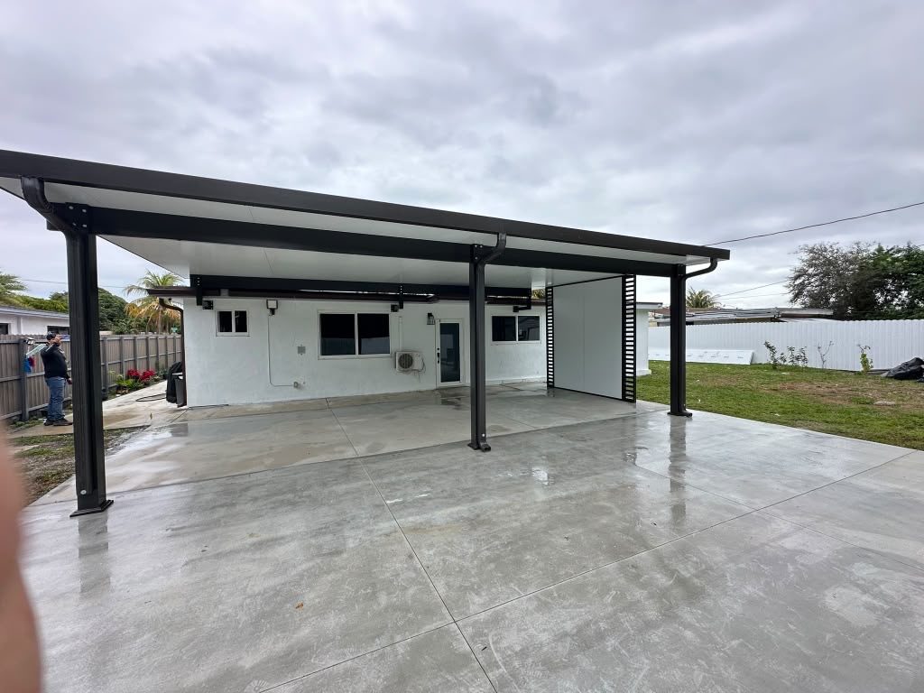 A newly poured concrete slab under a dark aluminium patio cover, still wet, with the garden fenced beyond
