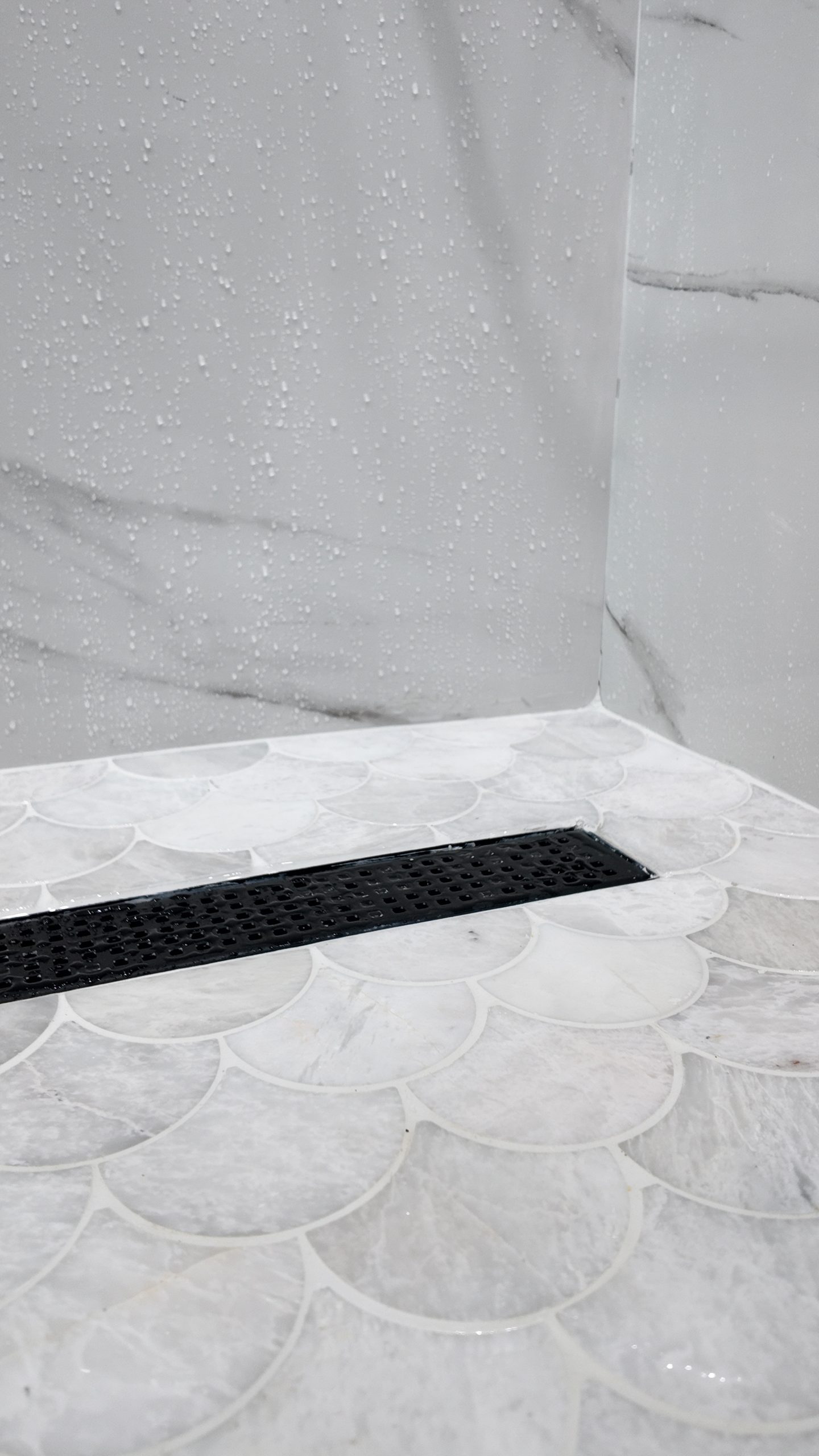 Shower floor laid in fish-scale mosaic, falling to a black linear drain set against large-format marble-effect wall tile