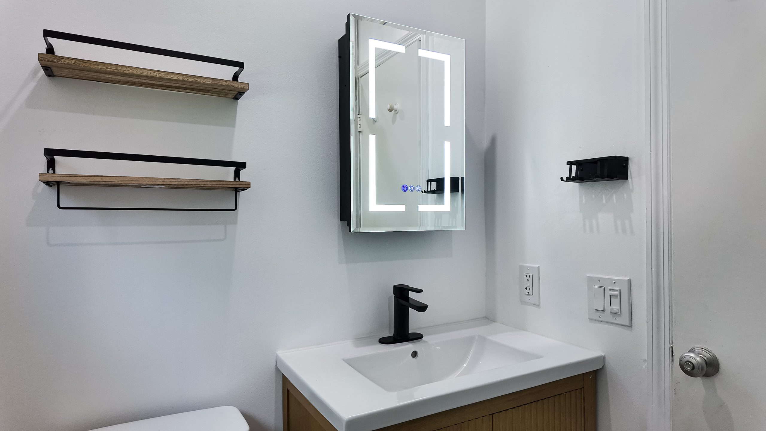 Completed small bathroom: wall-hung fluted vanity, lit mirror cabinet, open shelving and a window above the pan