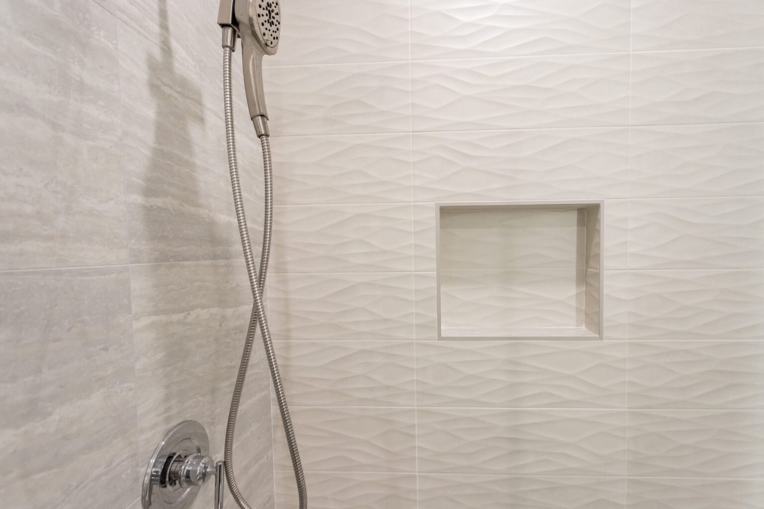 Shower wall detail: large-format wave-textured tile, a chrome handshower on a slide rail and a formed niche set into the tiling