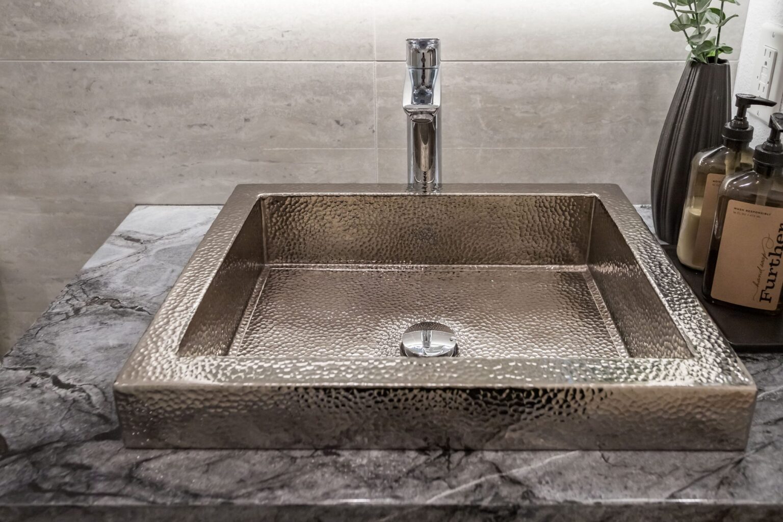 Hammered metal basin sitting on a grey-veined stone worktop with a tall chrome mixer and toiletry bottles beside it